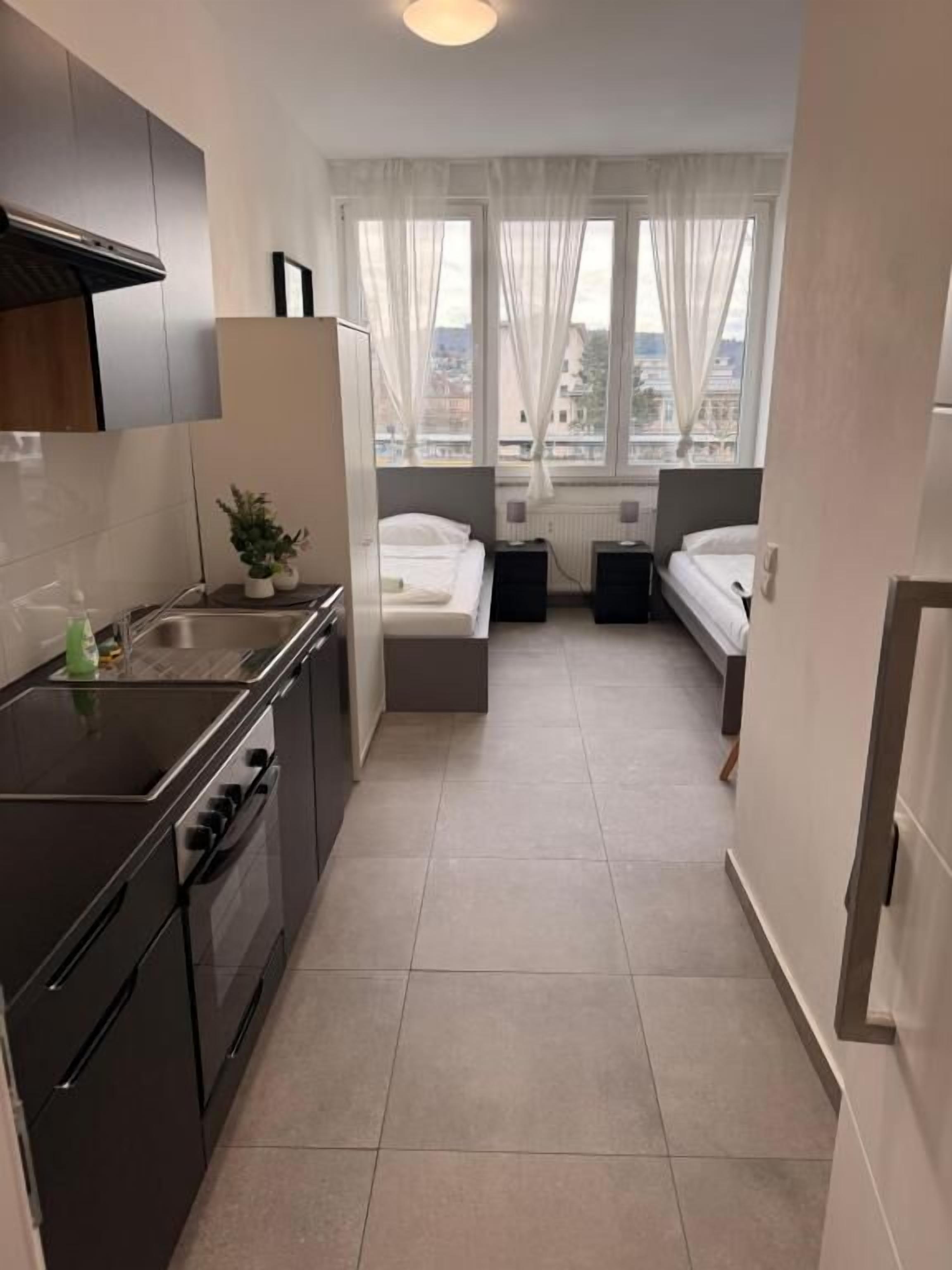 A compact hotel room with a kitchenette, a bed, and a view of the city through the window.