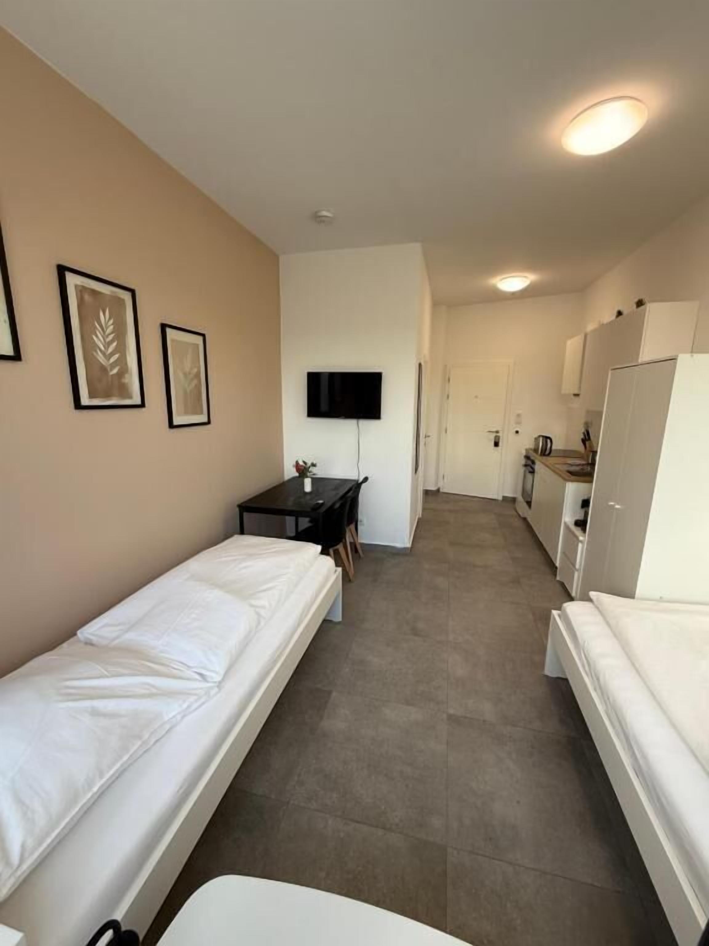 Ein Hotelzimmer mit zwei Einzelbetten, einem kleinen Tisch mit Stuhl, einem an der Wand befestigten Fernseher und einer Küchenzeile.
