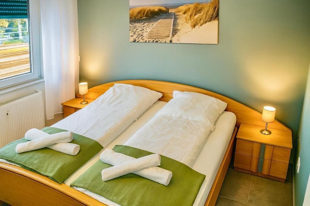 Cama doble con dos almohadas, una toalla verde y un cabecero de madera. Hay dos lámparas de noche y una ventana con persianas.