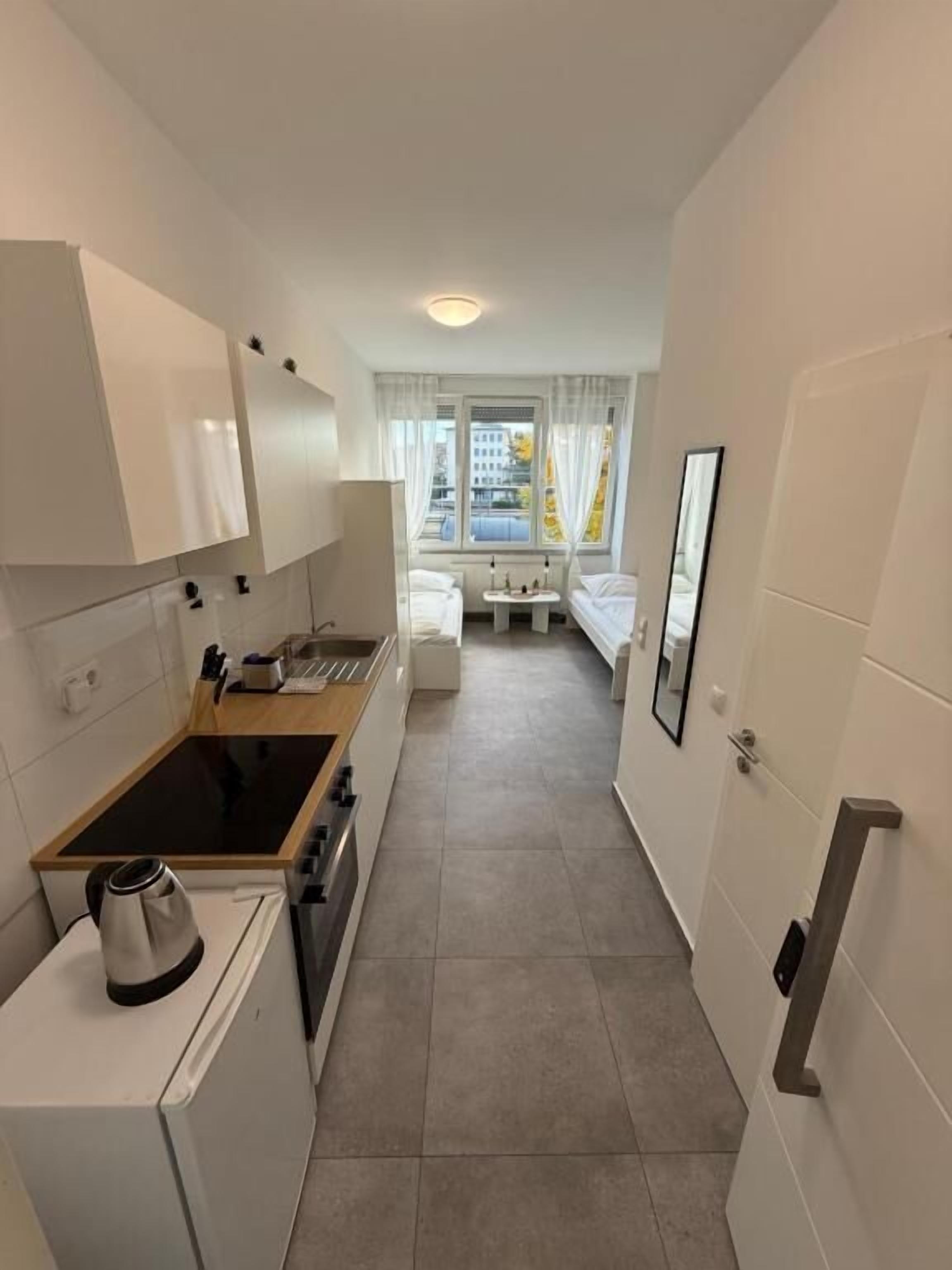 A narrow hotel corridor with a kitchenette on the left, a bed with a nightstand and a bedside table in the center, and a door on the right.