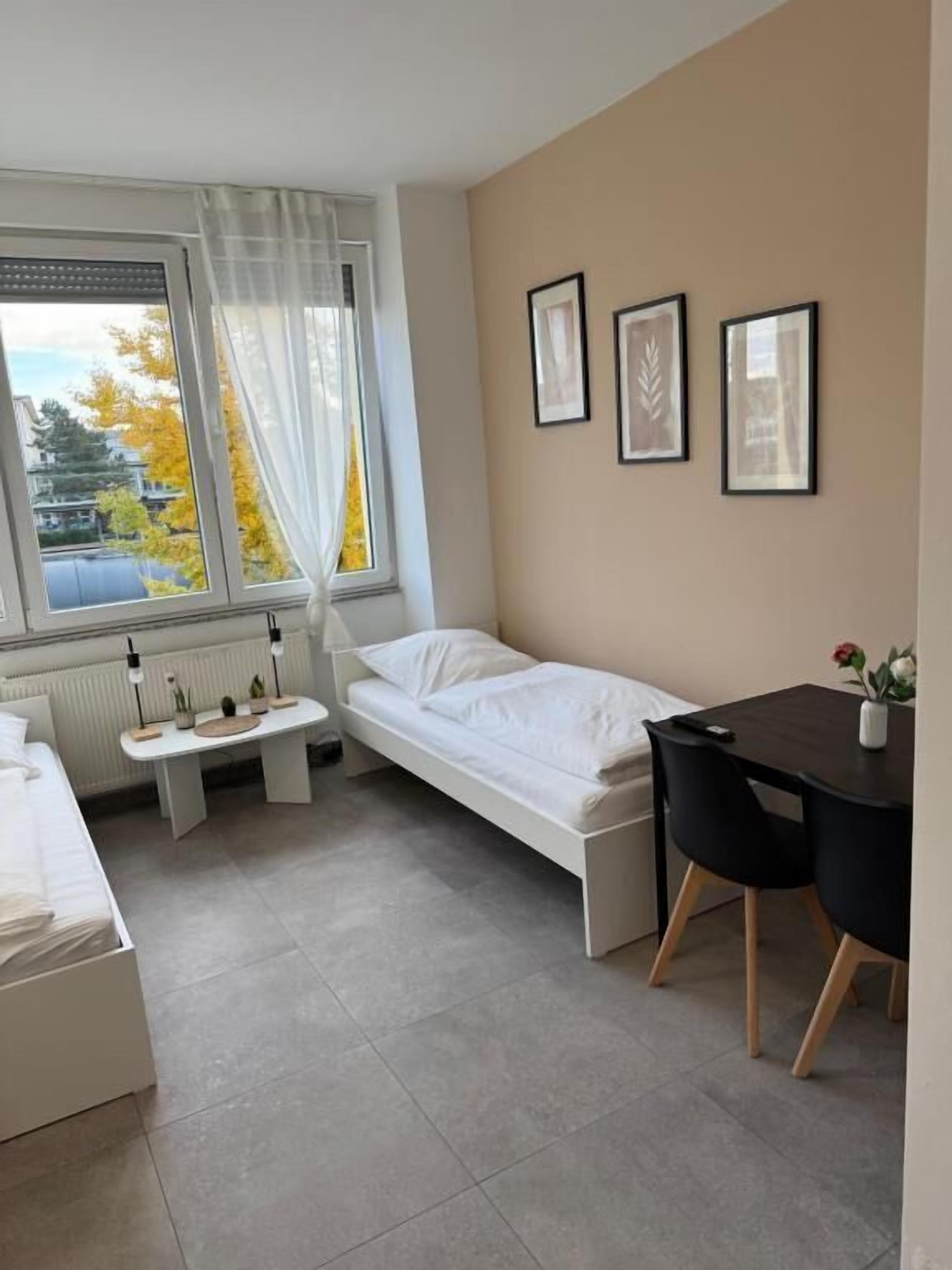 A compact room with a bed, a small table, a desk, and chairs. There are framed pictures on the wall.