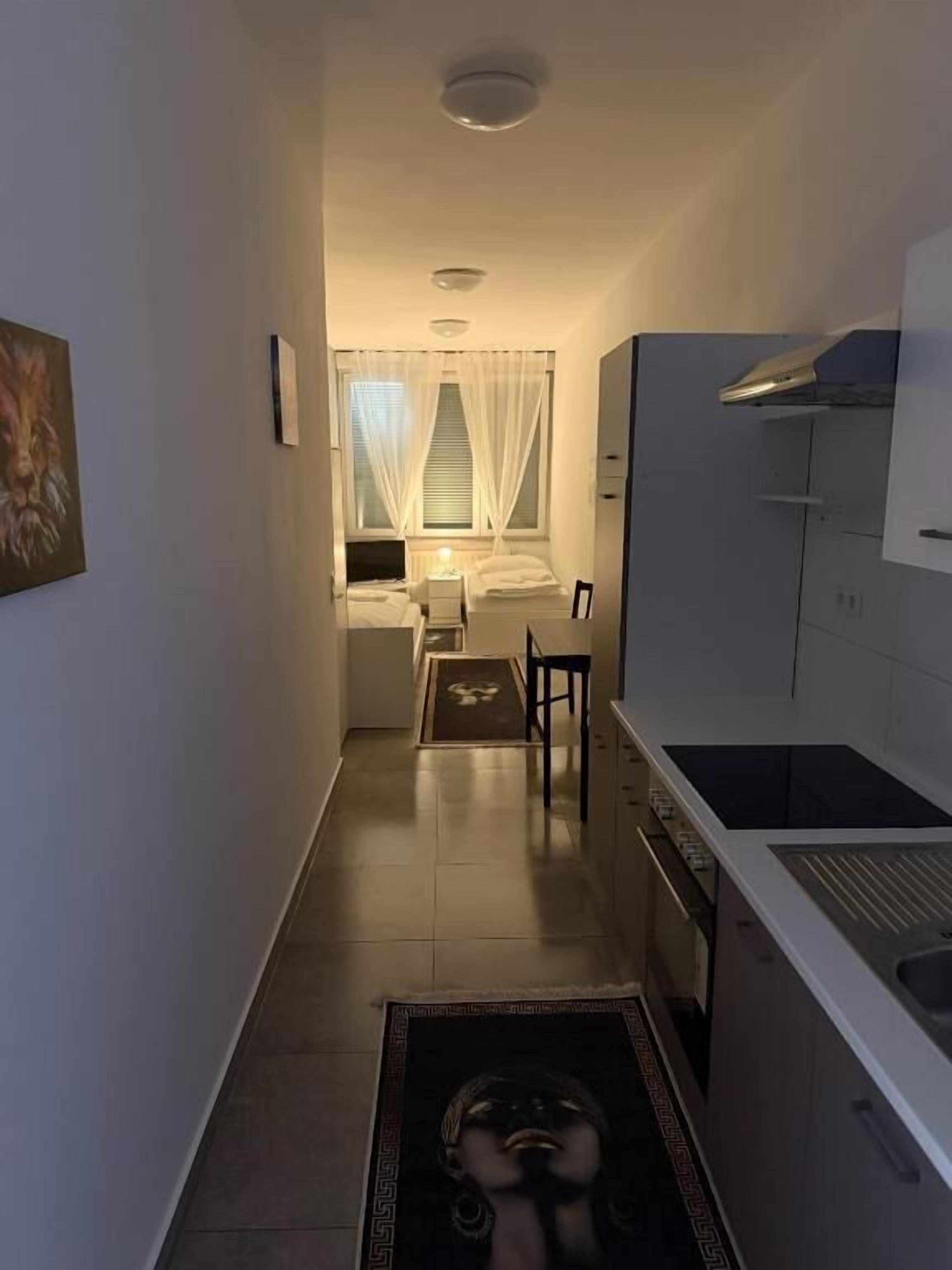 Een smalle gang die leidt naar een slaapkamer met een raam, een keuken met een fornuis, en een eettafel.