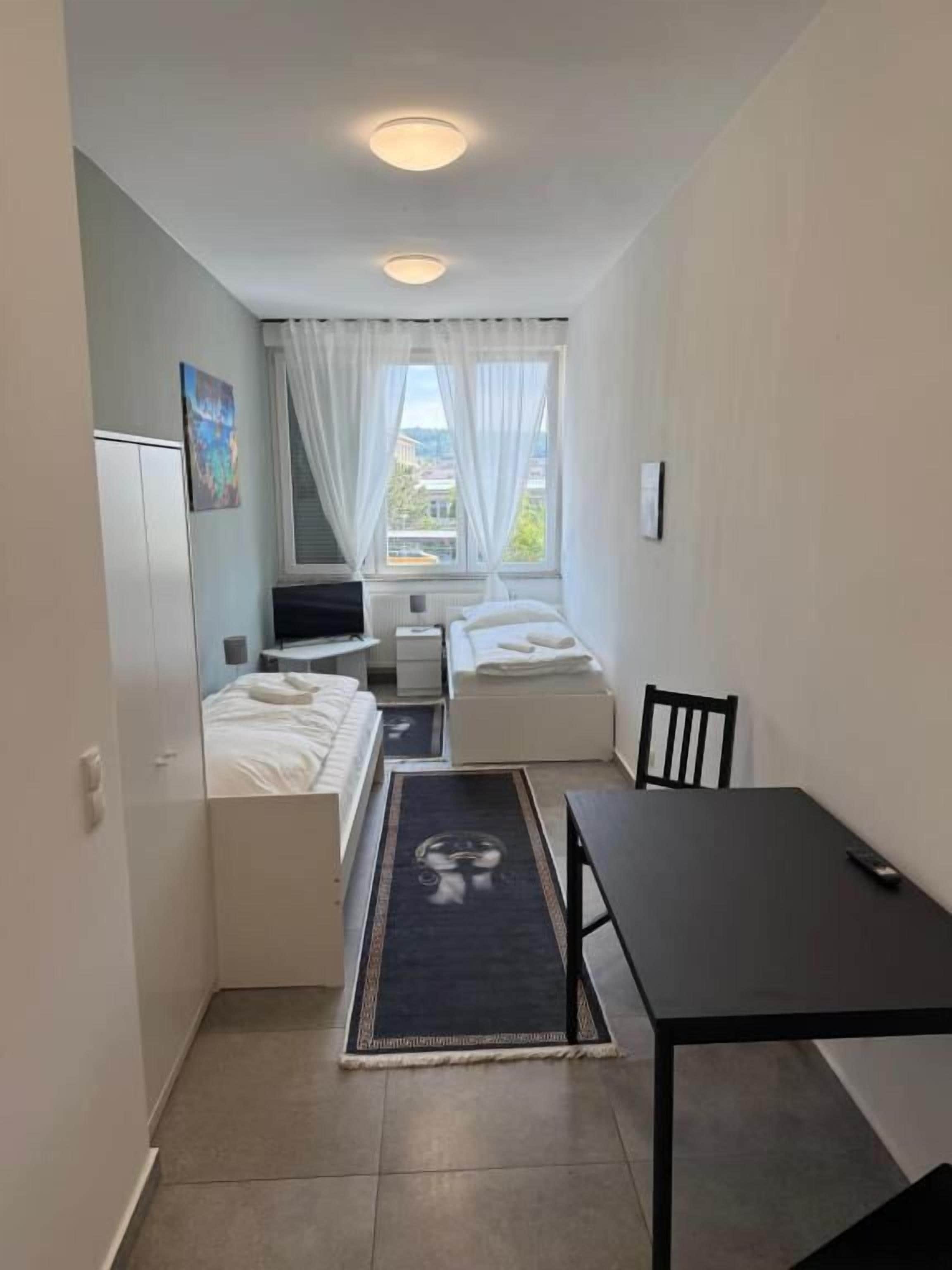 A compact room with a bed, a small table, a chair, a window with curtains, and a view of the outside.
