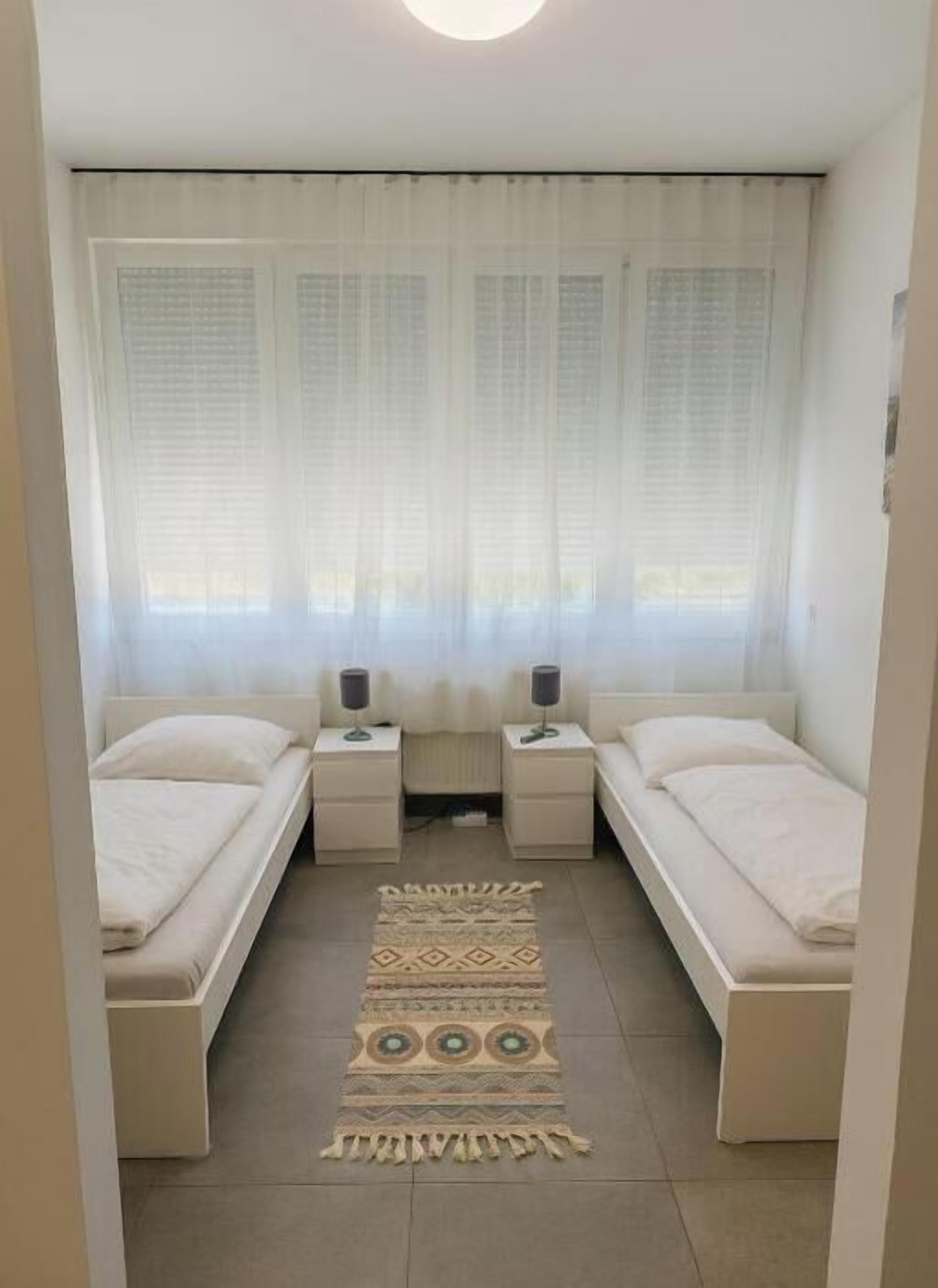 Un dormitorio con dos camas individuales, una mesita de noche con lámpara, una ventana con cortinas translúcidas y una alfombra estampada en el suelo.