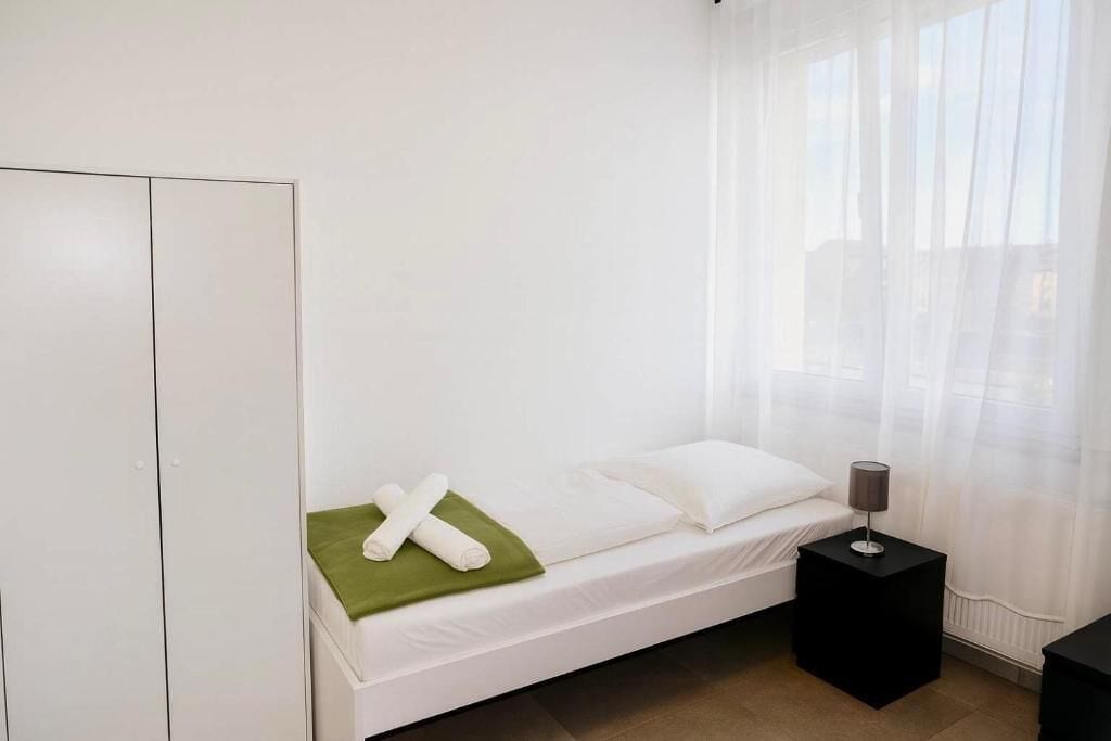 Uma cama de solteiro com roupa de cama branca, uma toalha verde e uma mesa de cabeceira preta com uma luminária.