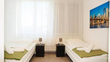 A hotel room with two beds, a cityscape painting on the wall, and a window with sheer curtains.