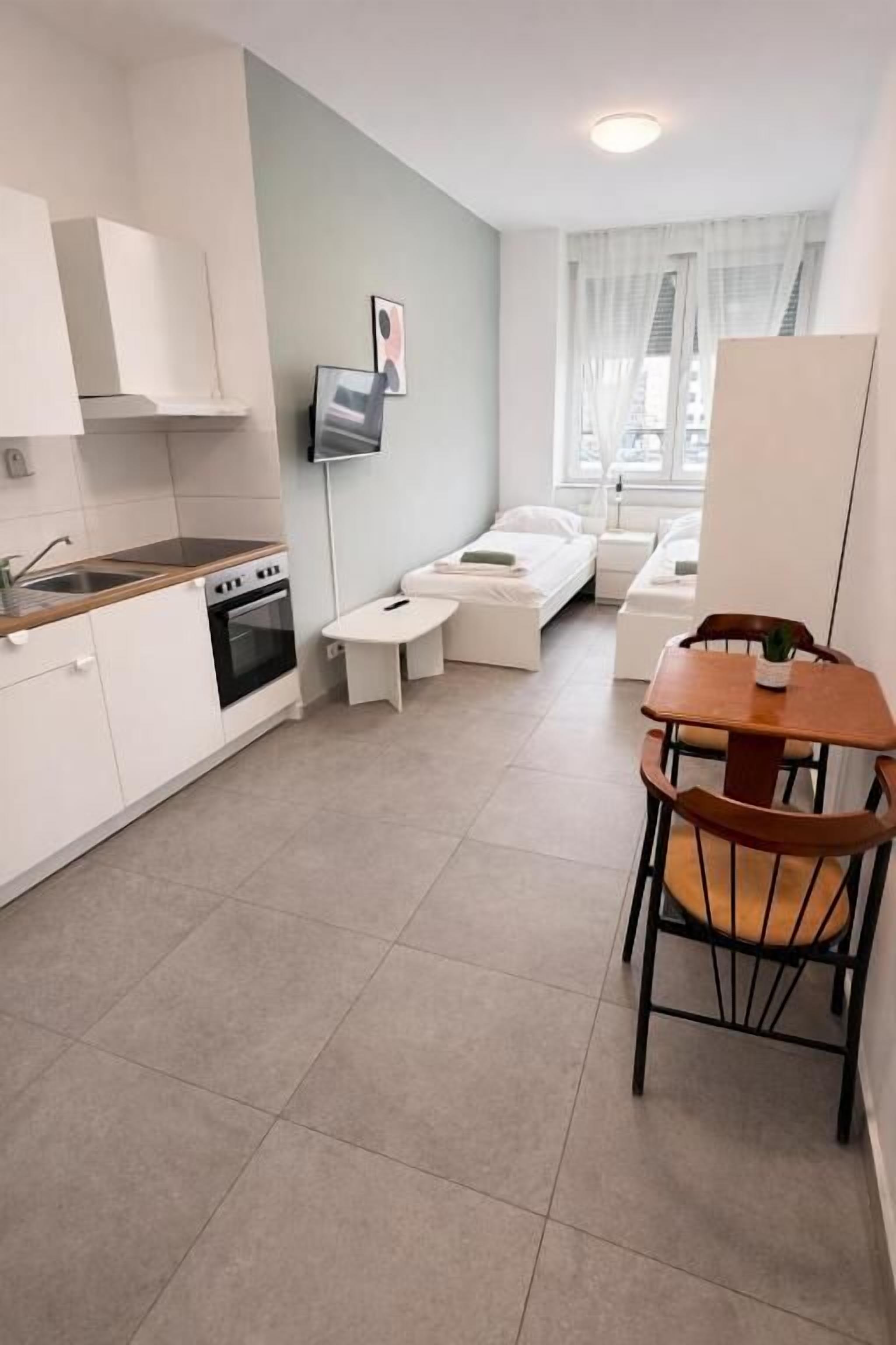 A compact hotel room with a kitchenette, a sofa, a dining table, and a TV mounted on the wall.