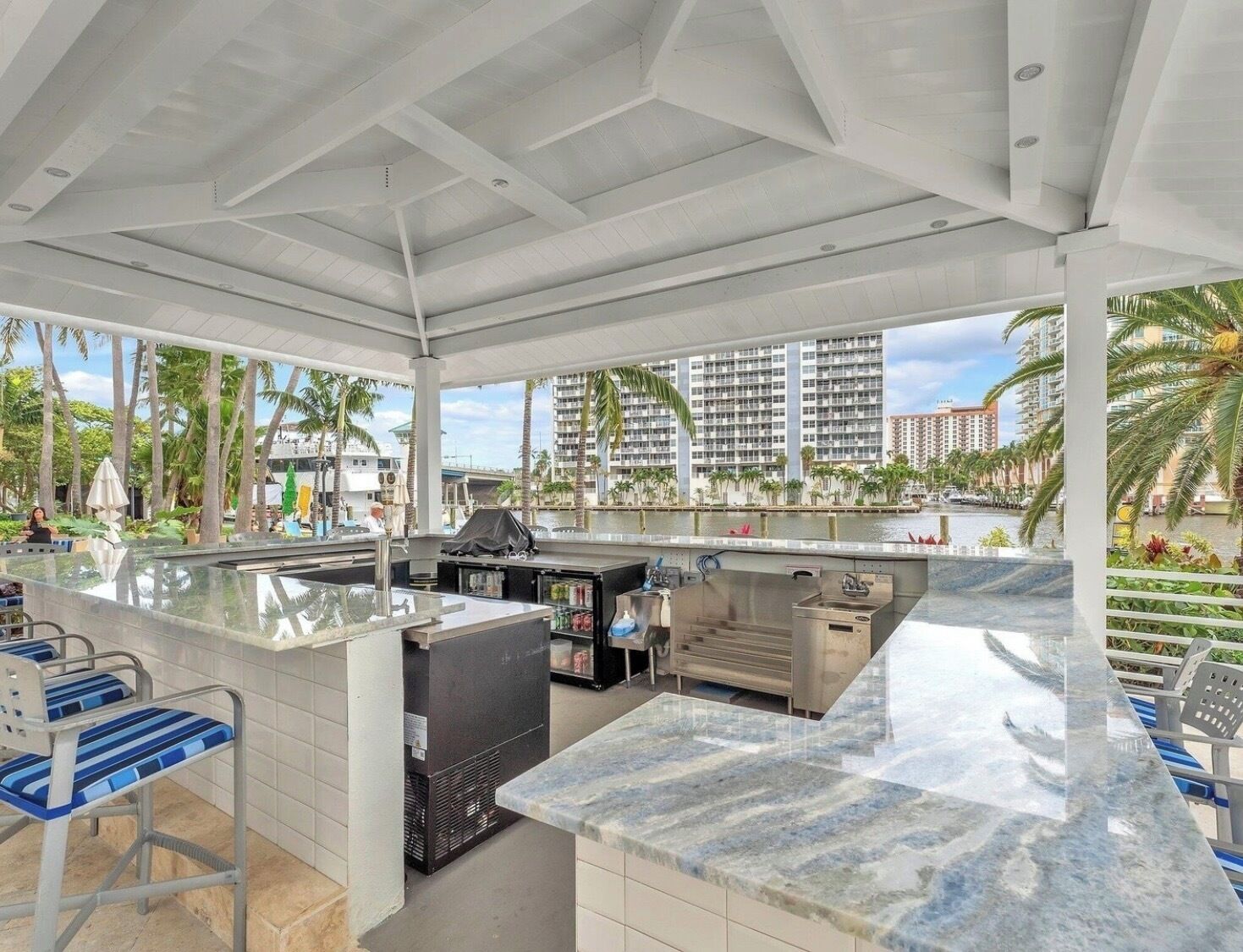 An outdoor bar area with a marble countertop, seating, and a view of a waterfront with buildings and palm trees.