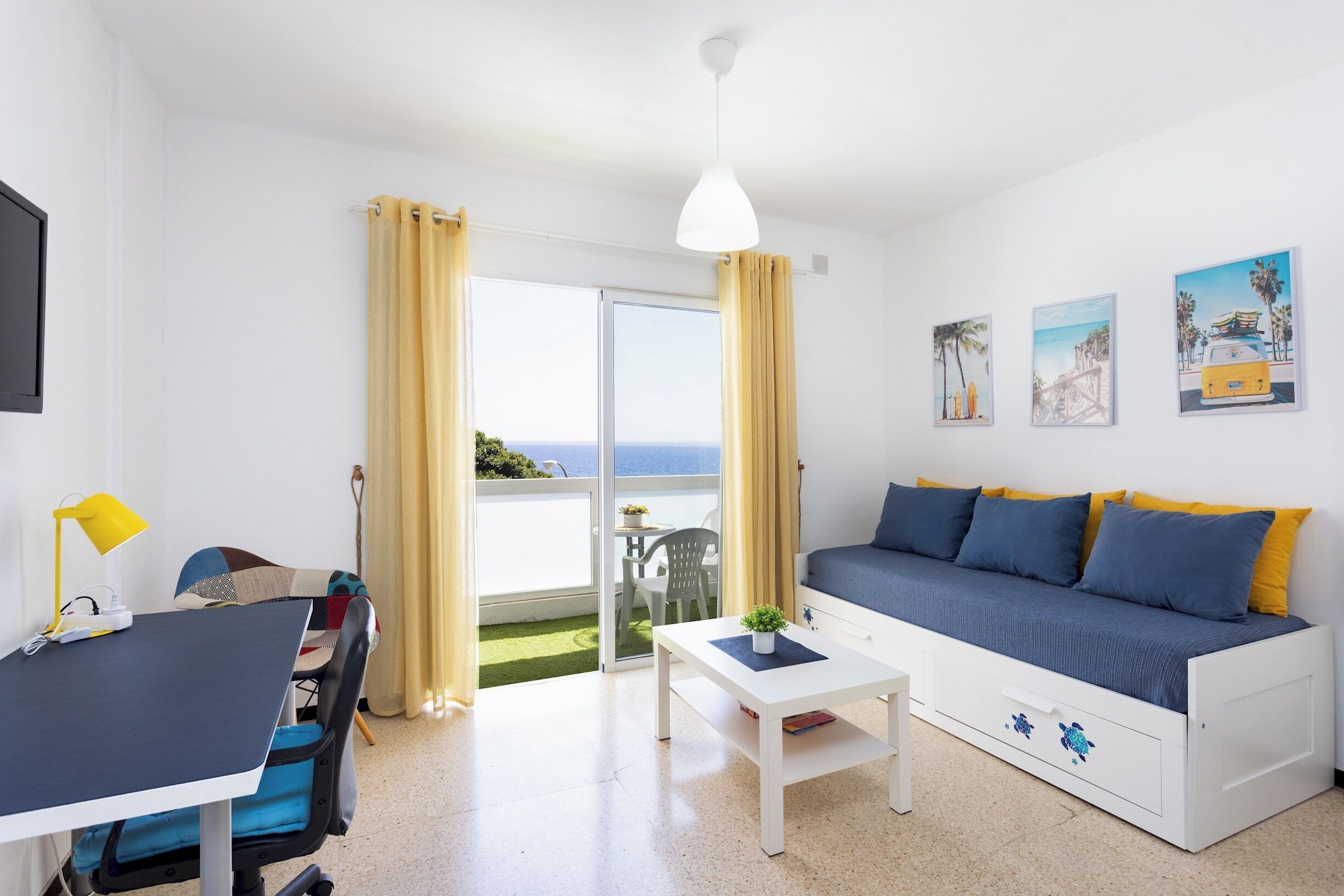 Un espace de vie compact comprenant un canapé, une petite table, un bureau avec une chaise et une vue sur la mer par une porte vitrée.
