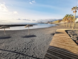 A beach with a wooden boardwalk, pebbles, and palm trees. - Home2Book Sea View Studio & Pool, Caletillas (Las Caletillas)