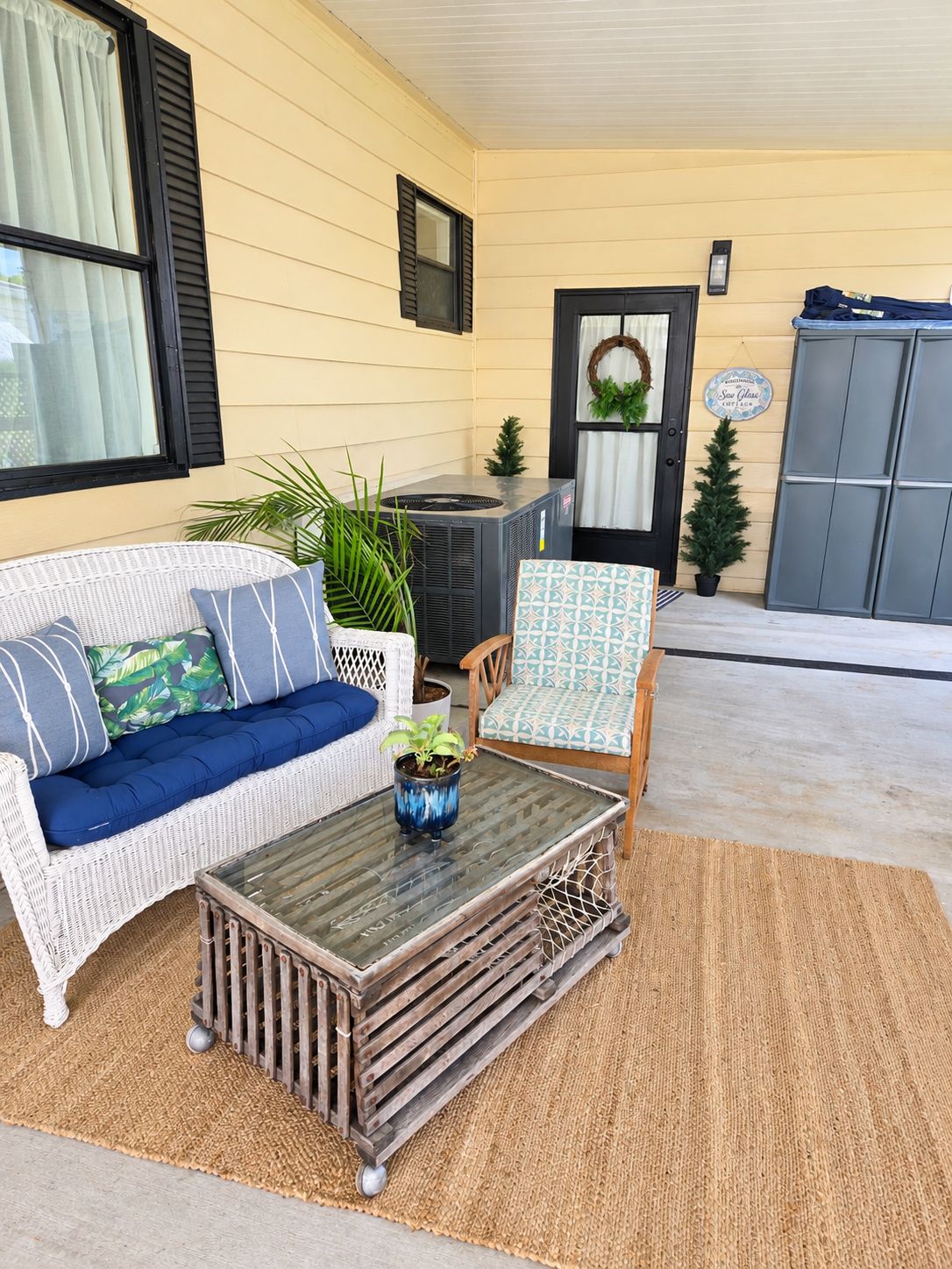 A covered porch with a wicker sofa, a wooden coffee table, and a chair with a patterned cushion.