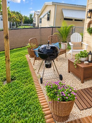 Un patio trasero con una parrilla, sillas de mimbre, una mesa de madera y plantas en macetas.