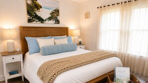 Una cama bien tendida con cabecera de madera, ropa de cama blanca y un edredón color beige. Hay dos mesitas de noche con lámparas y un pequeño cuadro enmarcado. Una cesta de mimbre con toallas enrolladas y una vela está en la mesita de noche.