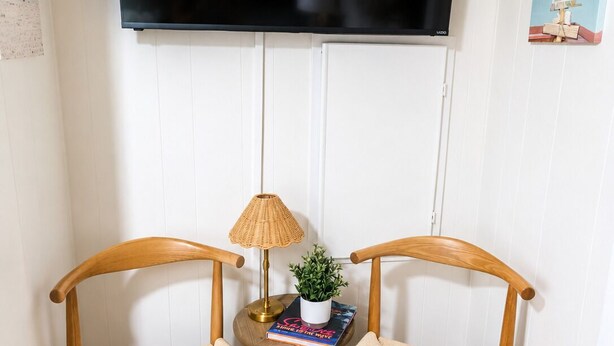 Una habitación con un televisor de pantalla plana montado en la pared, dos sillas de madera con asientos de mimbre, una mesita redonda con una lámpara y una planta en maceta, y un libro.
