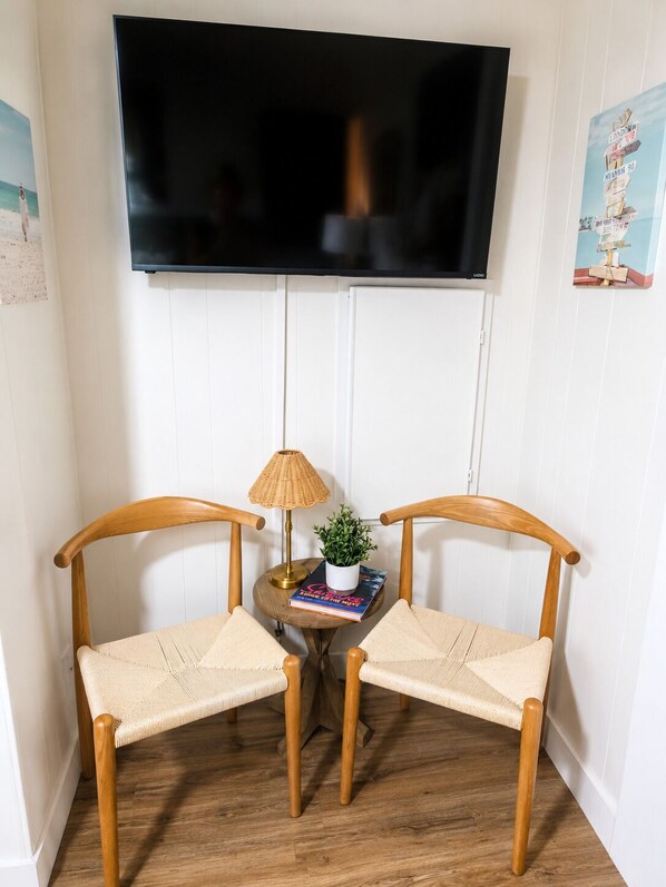 A room with a flat-screen TV mounted on the wall, two wooden chairs with woven seats, a small round table with a lamp and a potted plant, and a book.