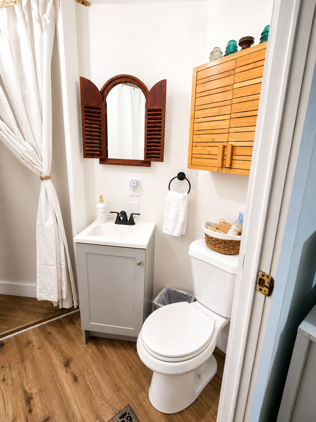 Un baño con lavamanos blanco, espejo con contraventanas de madera, inodoro y un pequeño cesto de basura.