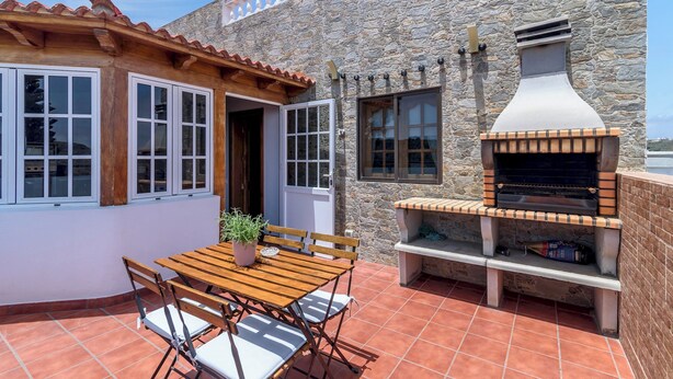 Een terras buiten, met een stenen muur, een houten pergola, een barbecue en een tafel met stoelen.