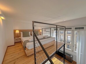 Une chambre moderne avec un lit superposé, un plancher en bois, une table de chevet avec une lampe et une fenêtre donnant sur des immeubles.
