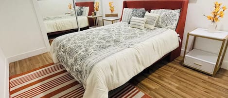 Ein Schlafzimmer mit einem großen Bett, einem Deckenventilator, einem Fenster mit Jalousien, einem Spiegel und einem kleinen Nachttisch mit Blumen in einer Vase.