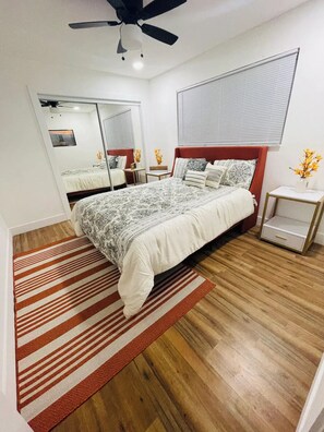 Ein Schlafzimmer mit einem großen Bett, einem Deckenventilator, einem Fenster mit Jalousien, einem Spiegel und einem kleinen Nachttisch mit Blumen in einer Vase.