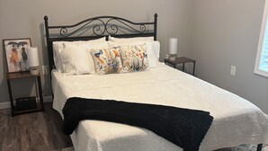 A bed with a black headboard, white bedding, and decorative pillows. There is a bedside table with a lamp and a framed picture. The room has a wooden floor and a patterned rug. - Cozy cottage with plenty of space in friendly Thaxton, MS. (Thaxton)