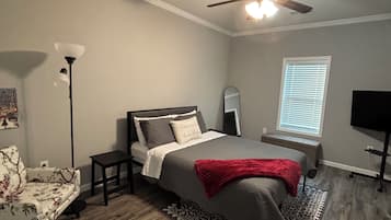 Une chambre à coucher comprenant un lit, un ventilateur de plafond, une lampe sur pied, un téléviseur et une fenêtre avec des stores.