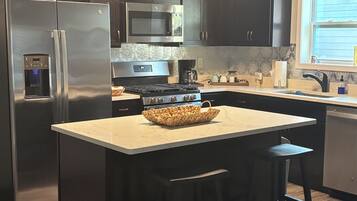 Une cuisine moderne avec des meubles de couleur sombre, des appareils en acier inoxydable, un îlot central doté de tabourets et une fenêtre laissant entrer la lumière naturelle.