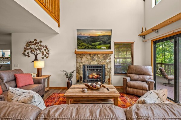 Ein Wohnzimmer mit Kamin aus Stein, Ledersofas, einem Couchtisch mit Dekoration und einem großen Fenster mit Blick auf Grünflächen.