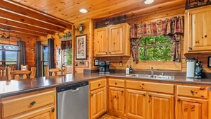 A wooden cabin interior with a kitchen area featuring wooden cabinets, a dishwasher, and a window with a view of greenery. - Incredible MTN Views*Hot Tub + Game Room*Fireplace (Pigeon Forge)
