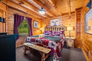 A wooden cabin bedroom with a bed, bench, and a window with curtains. - Incredible MTN Views*Hot Tub + Game Room*Fireplace (Pigeon Forge)