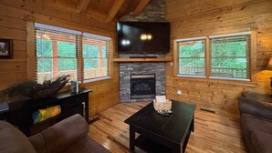 Interior acogedor de una cabaña con chimenea, paredes de madera, un televisor de pantalla plana montado encima y una zona de estar cómoda con un sofá y una mesita de centro.