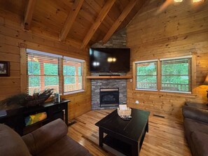 Ein gemütliches Blockhaus-Interieur mit Kamin, Holzwänden, einem flachbildfernseher, der darüber angebracht ist, und einer komfortablen Sitzecke mit Sofa und Couchtisch.