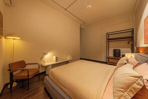 A hotel room with a bed, a desk with a chair, a shelf with books, and a wall-mounted rack with a suitcase. - The Cozy Hideout: Studio Inside a Charming Cafe (Cau Ong Lanh)
