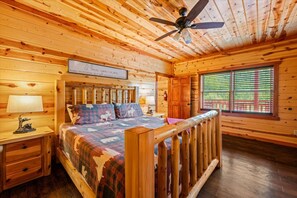 Une chambre dans un chalet en bois, comprenant un lit, une table de chevet, une lampe, un ventilateur de plafond et une fenêtre avec des stores.