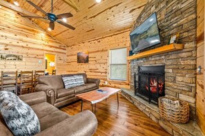 Ein gemütliches Blockhaus-Interieur mit Kamin aus Stein, Holzwänden, einer Ledersofa und einem Deckenventilator.