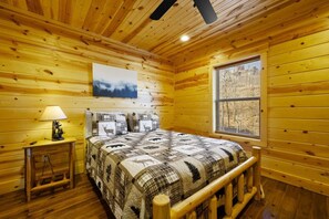 A wooden cabin bedroom with a bed, a nightstand, a lamp, a window, and a painting on the wall. - New! 3BR w/Hot Tub*Game Room*Comm Pools*Fireplace (Gatlinburg)