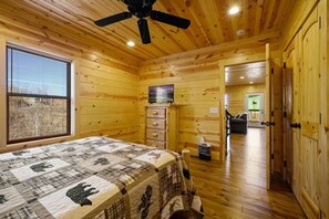Un dormitorio de cabaña de madera con cama, ventilador de techo, ventana con persianas y puerta que da a otra habitación.