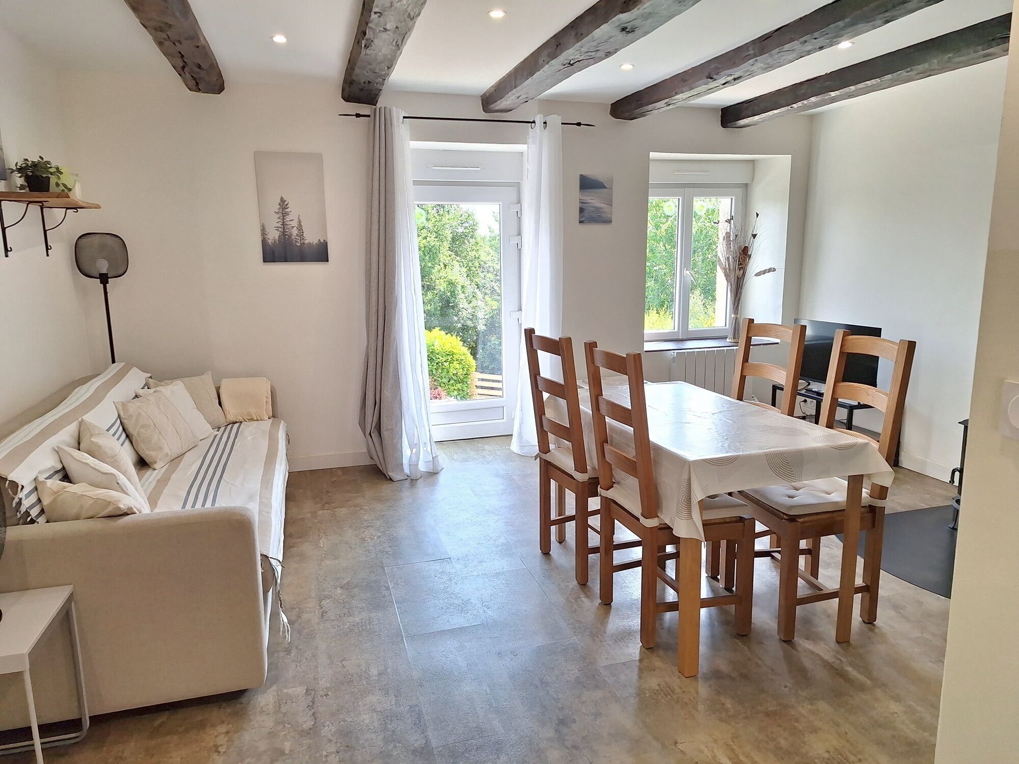 A room with a dining table and chairs, a sofa, a bed, and a view of greenery outside.
