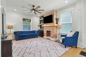A living room with a blue sofa, a fireplace, a ceiling fan, and a large window with blinds. - Luxurious curated high end oasis (Cincinnati)