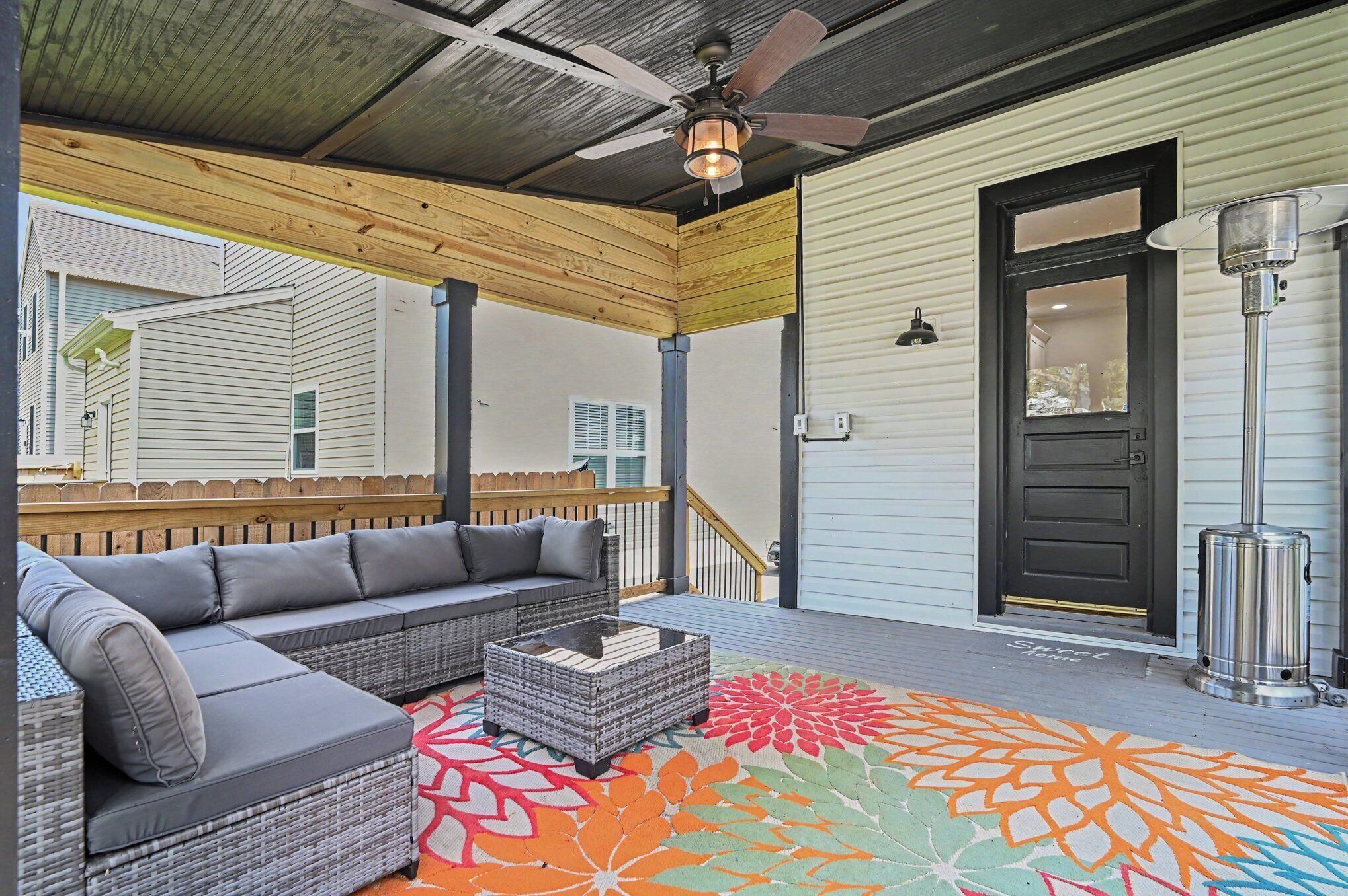 A covered porch with a gray sectional sofa, wicker coffee table, and a colorful rug.