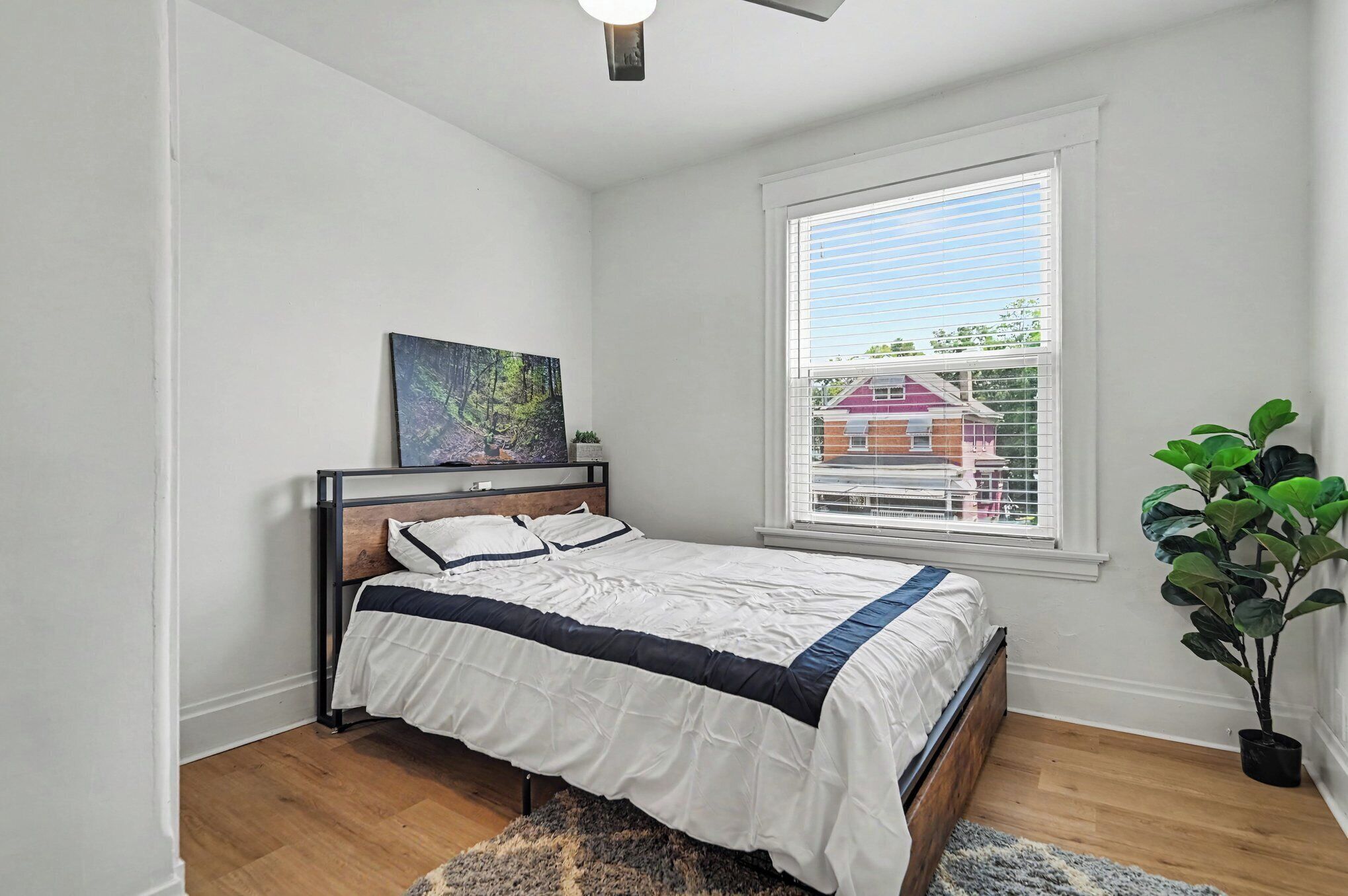 Une chambre à coucher avec un lit, un tableau au mur, une fenêtre avec des stores et une plante en pot.