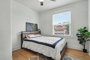 Une chambre à coucher avec un lit, un tableau au mur, une fenêtre avec des stores et une plante en pot.