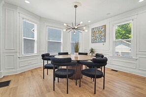 Uma sala de jantar com uma mesa redonda e seis cadeiras, paredes brancas e um lustre.