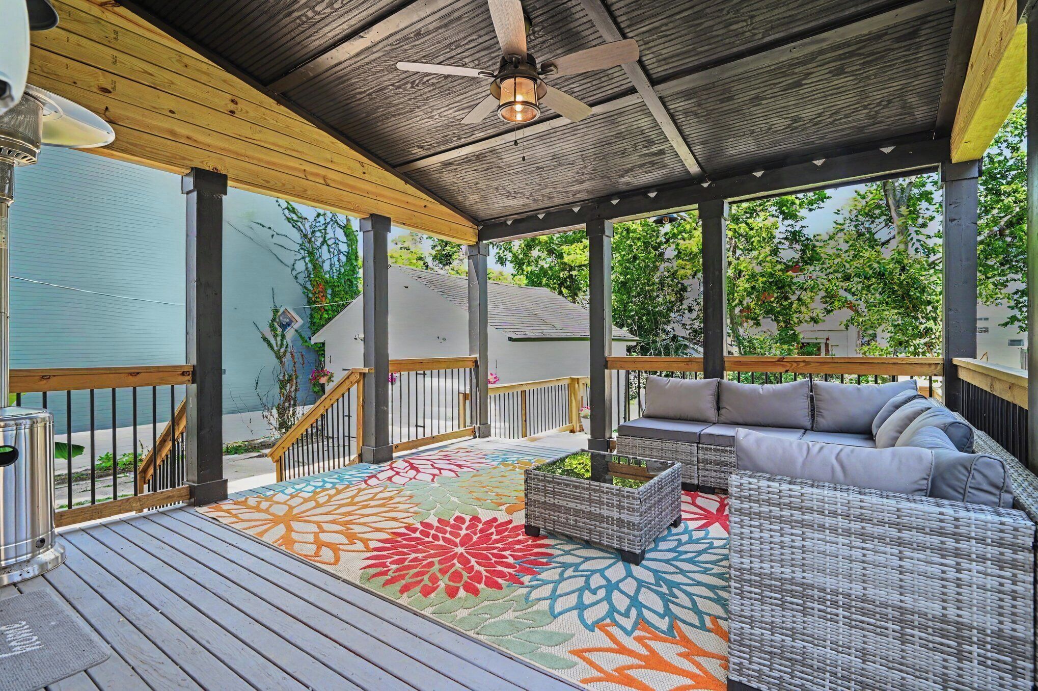 A covered outdoor area with a wooden deck, a ceiling fan, a colorful rug, and outdoor furniture.