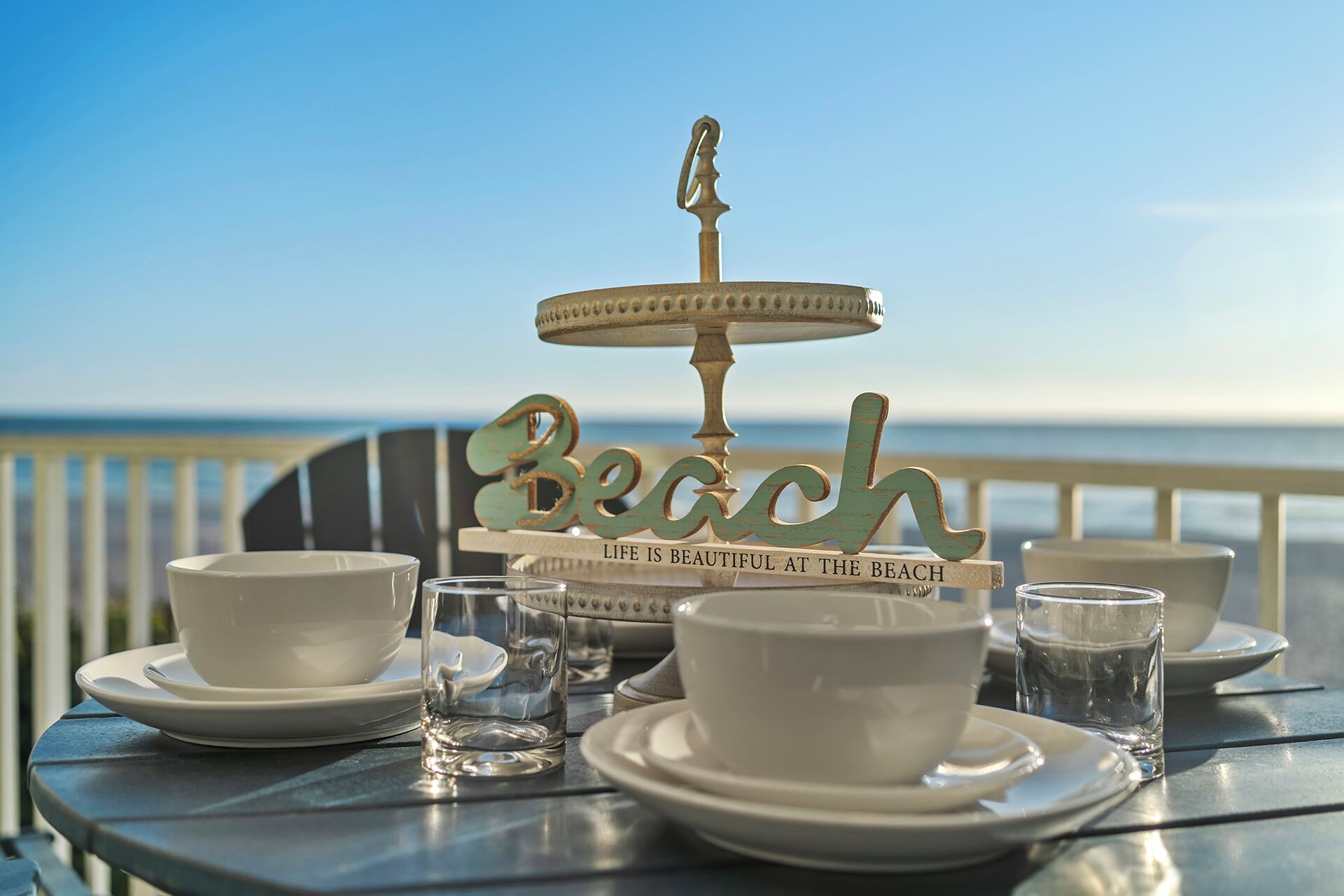 Una mesa dispuesta con tazas y platillos blancos, un vaso y un letrero decorativo con la palabra 'Beach' y una cita.