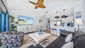 A modern living room with a flat-screen TV, a dining area, and a kitchen with white cabinets and stainless steel appliances. - Top-Floor Gulf Front · Sunset Balcony · Heated Pool (Holmes Beach)