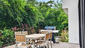 Un patio al aire libre con mesa y sillas de madera, una parrilla y abundante vegetación.