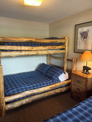Ein Stockbett mit Holzrahmen, einer blauen Karo-Matratze und einem kleinen Nachttisch mit Schubladen.