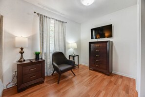A room with a wooden floor, a dark-colored chair, a small table with a lamp, a cabinet with drawers, and a television mounted on the wall. - I129 - 3BD Pool Retreat | Fire Pit & Backyard | Irving (Irving)