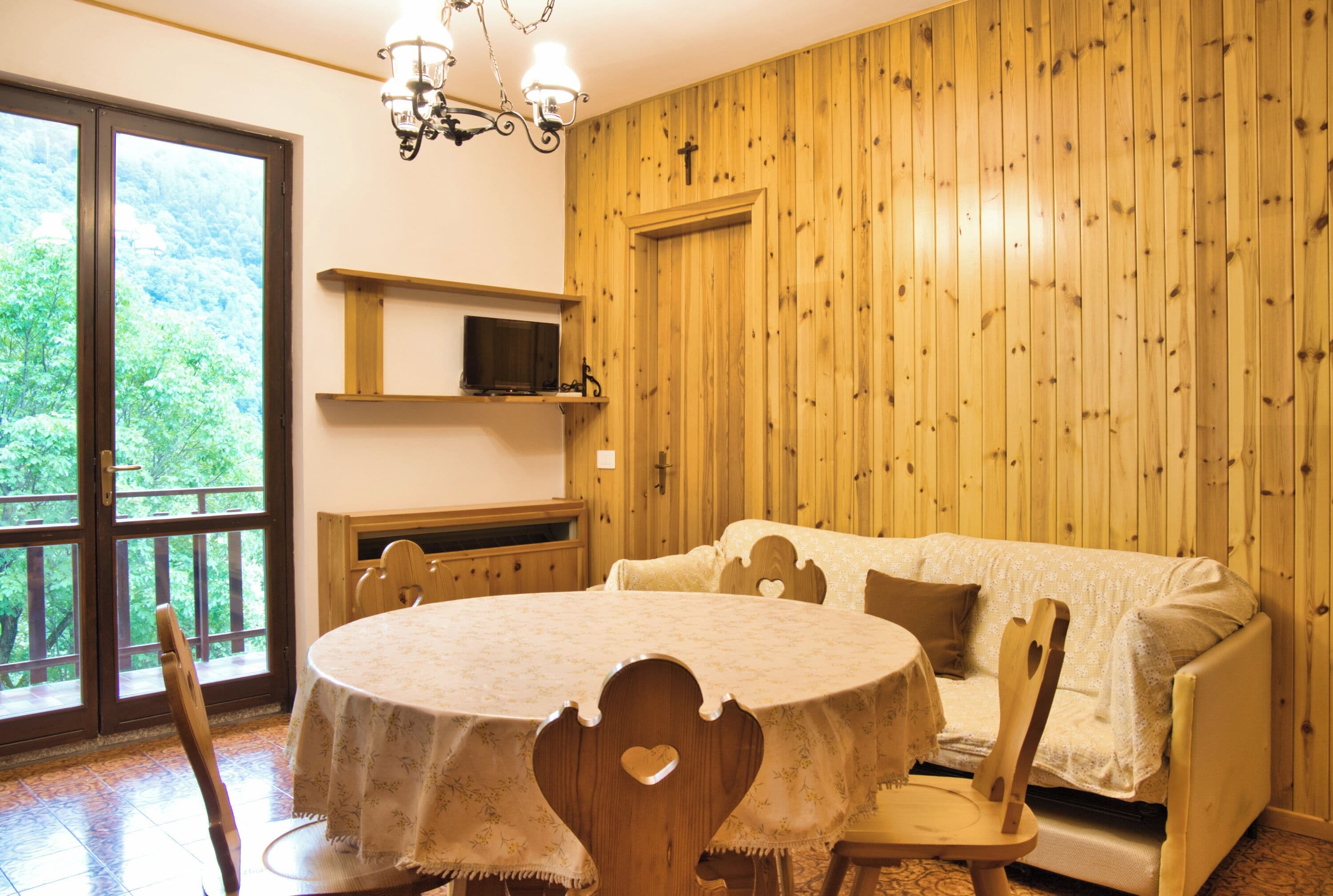 Een gezellige kamer met een ronde tafel, stoelen, een bank, een televisie op een plank en een houten wand met een kruis.